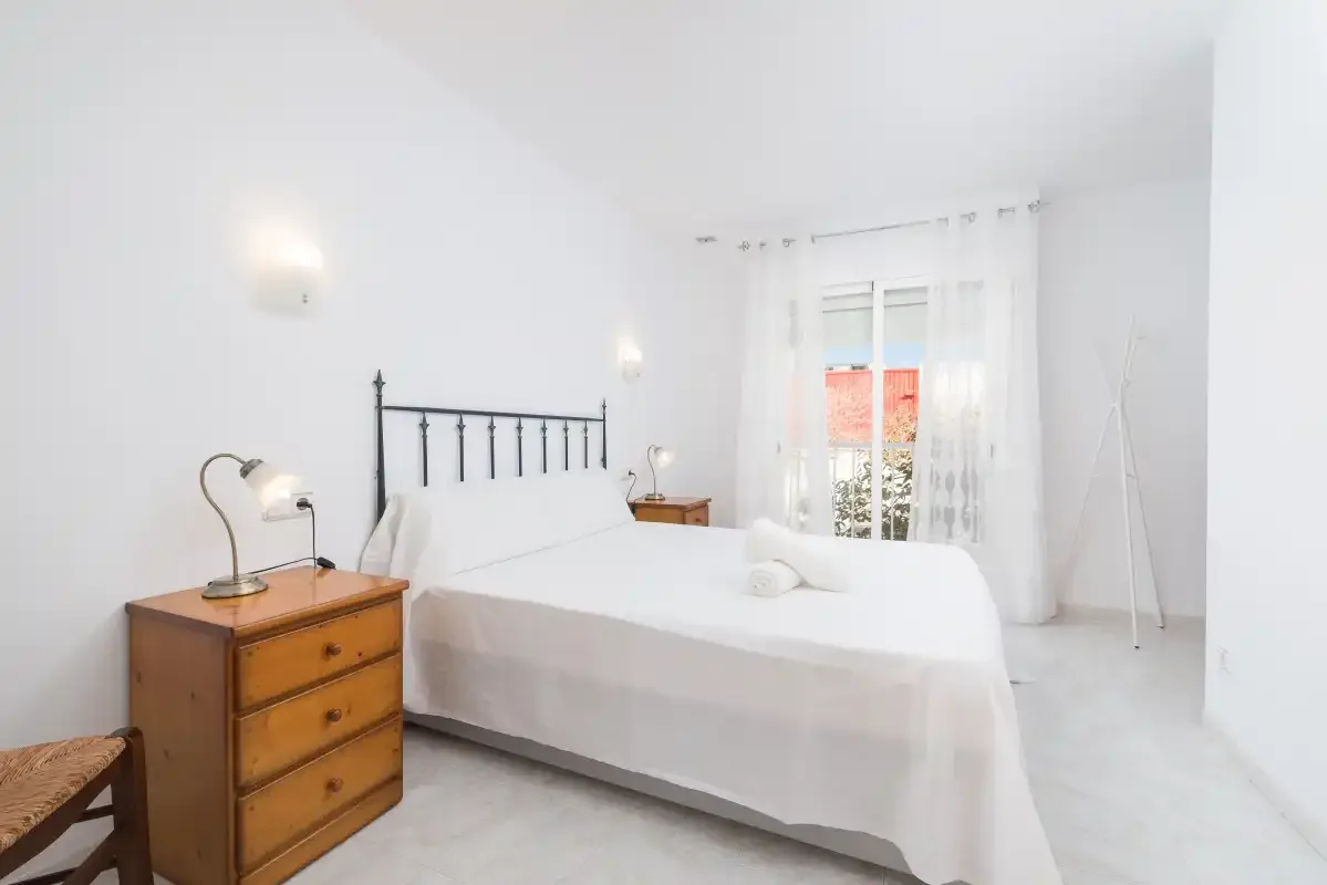 A bright bedroom with a bed, wooden dresser, and window with sheer curtains.
