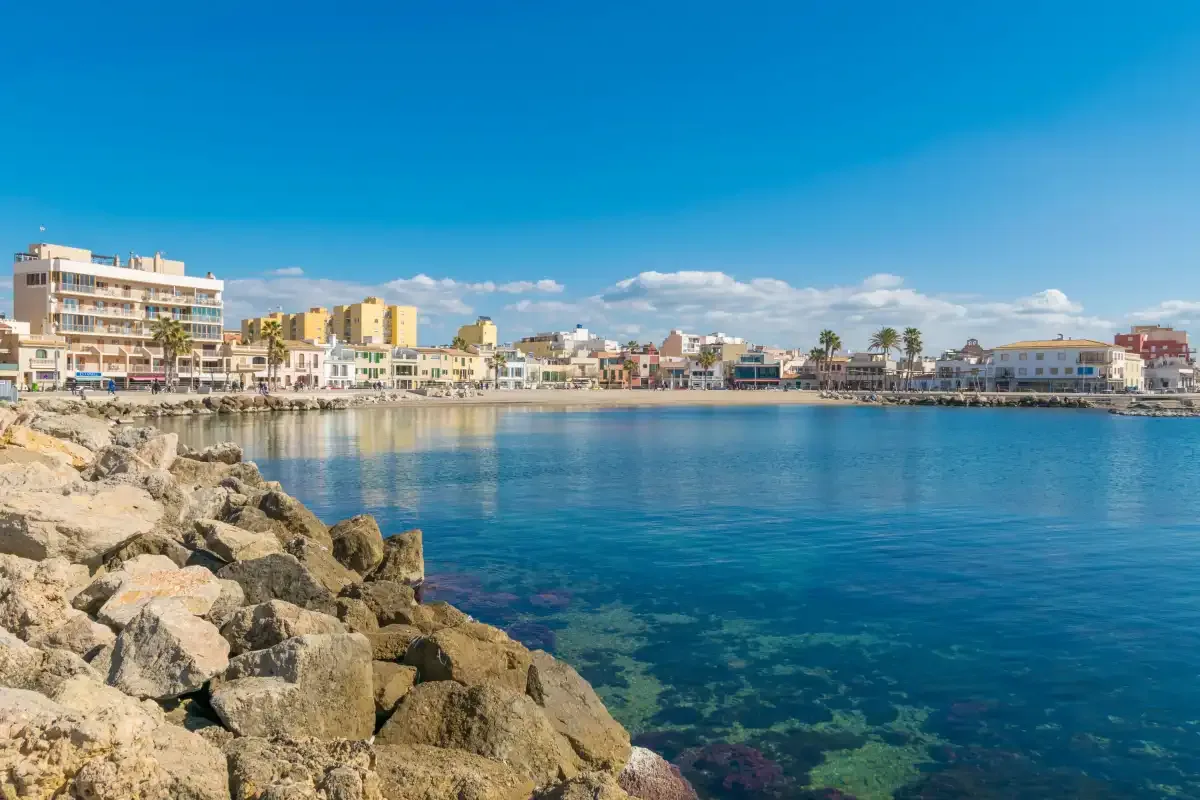 A coastal waterfront with rocky shore, calm blue water, and buildings in the distance.