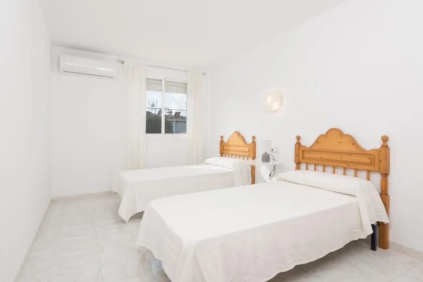 A bright bedroom with two beds, a window, and a wall-mounted air conditioner.