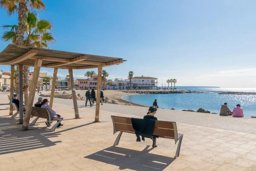 A sunny seaside promenade with shaded seating and people beside the water.