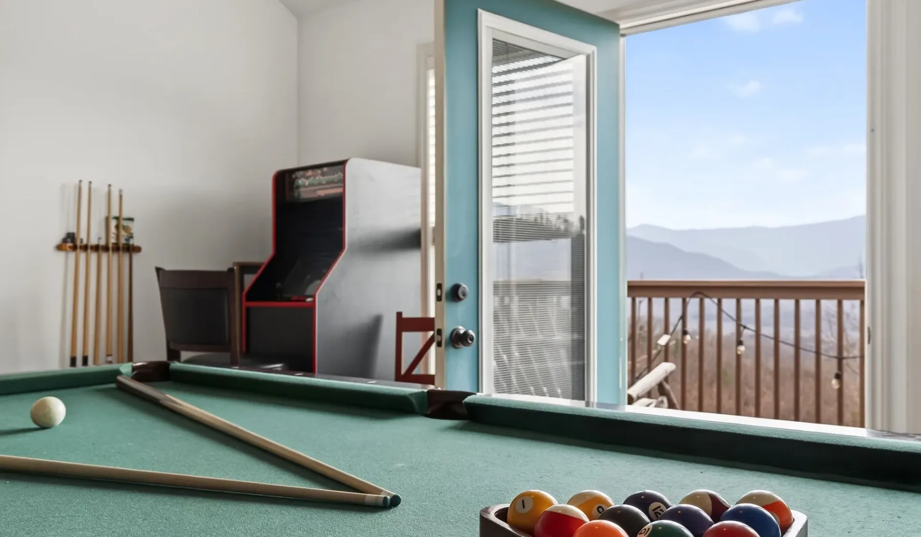 Una sala de juegos con mesa de billar, máquina recreativa y puerta abierta al balcón.