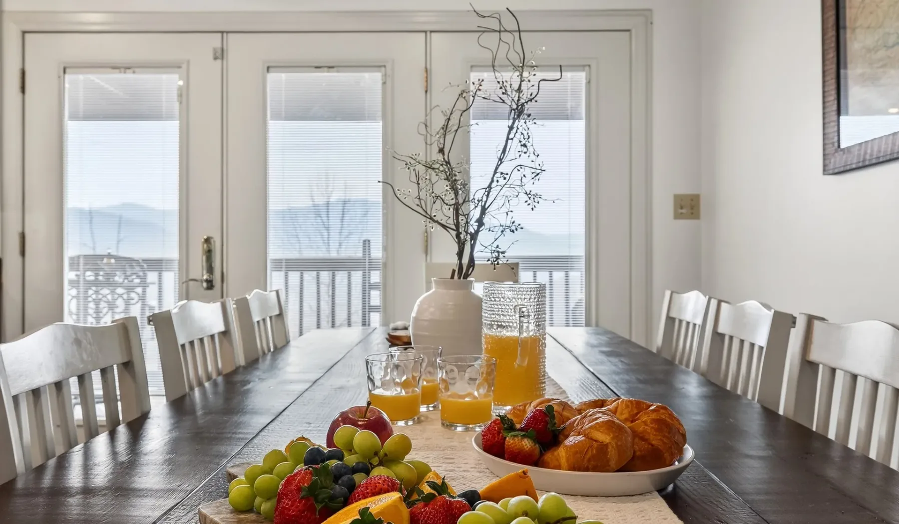 Una mesa de comedor tiene fruta, jugo y pasteles en una habitación luminosa.