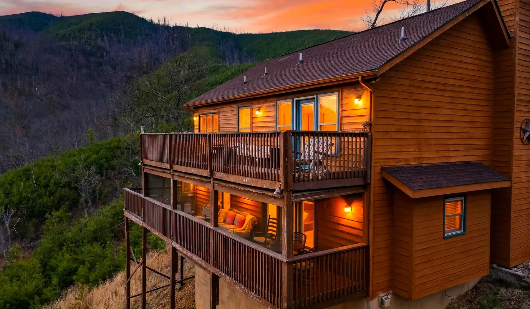 Una cabaña de madera con balcones iluminados domina un paisaje montañoso boscoso al atardecer.
