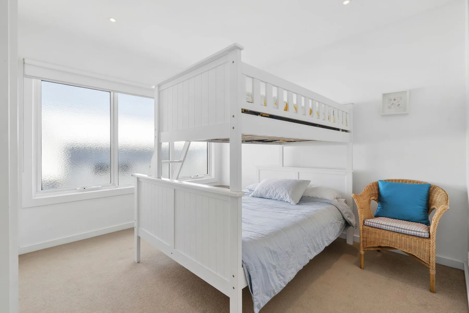 A bright bedroom has a white bunk bed and a wicker chair by the window.