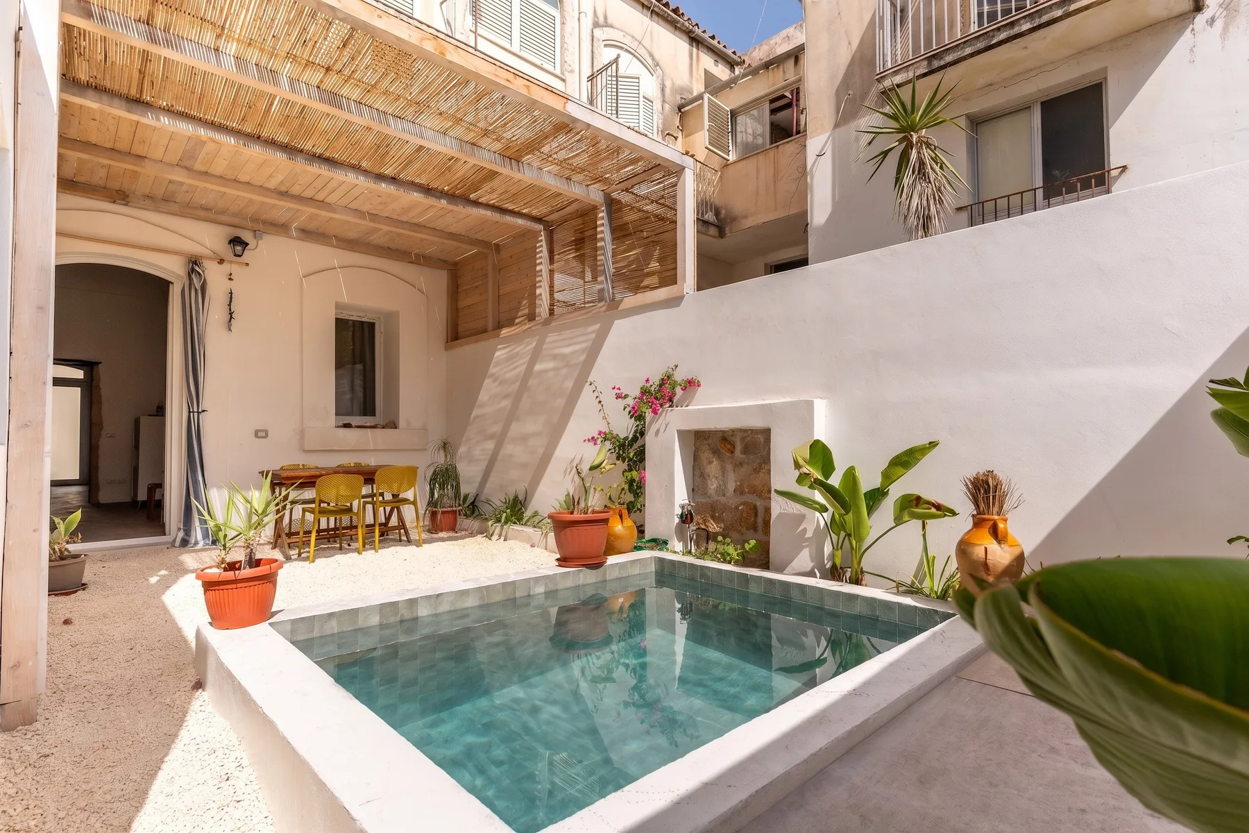 Una pequeña piscina de patio está junto a una zona de comedor sombreada y plantas en macetas.