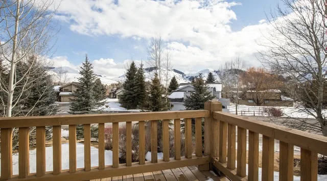 Una terraza de madera da a casas cubiertas de nieve, árboles perennes y montañas lejanas.