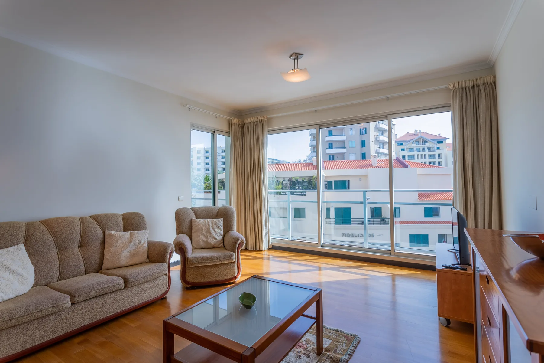 A bright living room with seating, a glass coffee table, and large windows.