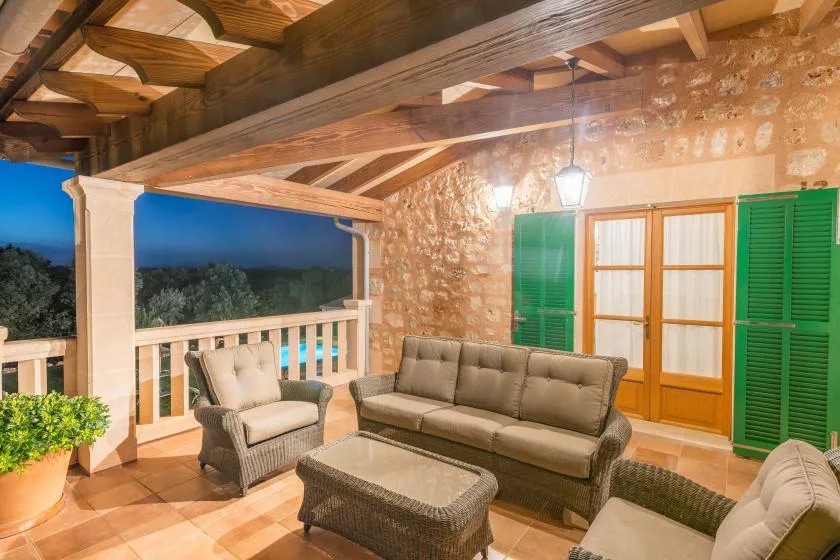 Una terraza cubierta con asientos de mimbre, vigas de madera y contraventanas verdes.