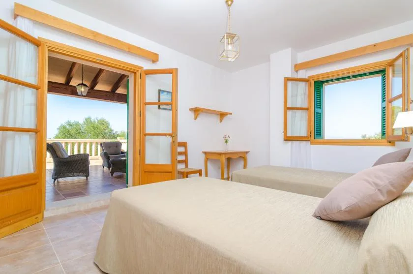Un dormitorio luminoso con dos camas, ventanas de marco de madera y una puerta abierta a una terraza.