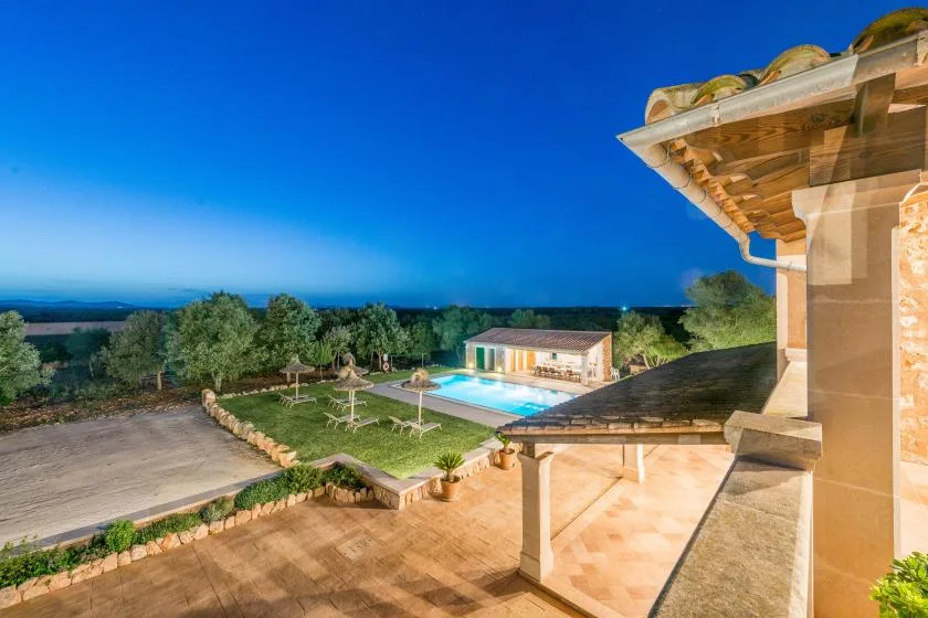 Una terraza de piedra da a una piscina y a un edificio bajo bajo un cielo despejado al anochecer.