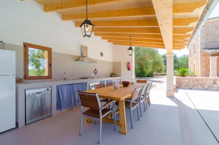 Una zona exterior cubierta para comer tiene una mesa larga bajo un techo con vigas de madera.
