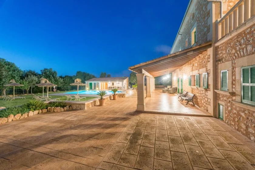 Un patio de piedra junto a un edificio da a una piscina iluminada y un jardín.