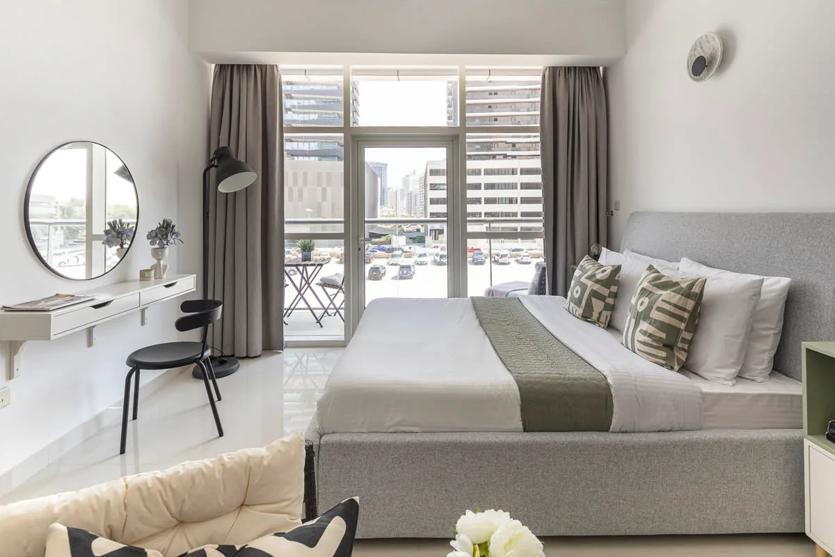 Un dormitorio luminoso con una cama junto a ventanas altas y un pequeño escritorio.