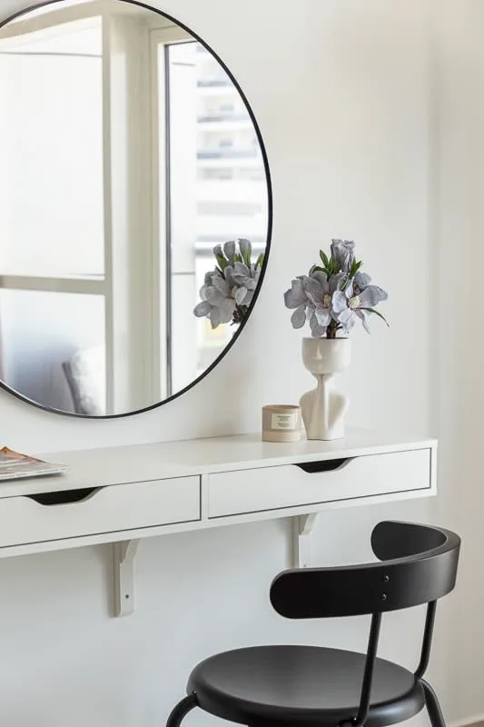 Una habitación luminosa con un escritorio blanco, un espejo redondo y una silla negra.
