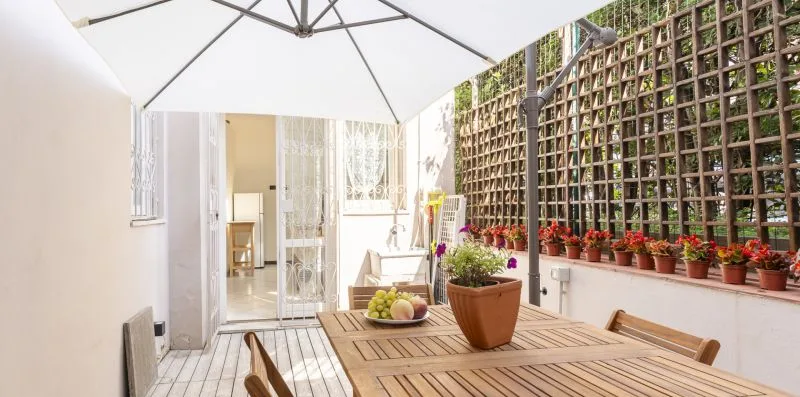 An outdoor patio has a wooden table, potted flowers, and a large umbrella.