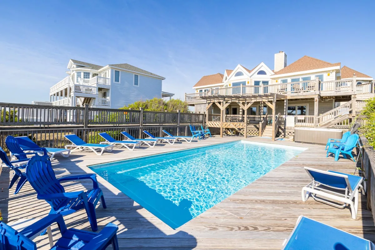 A swimming pool sits on a wooden deck beside coastal-style houses.