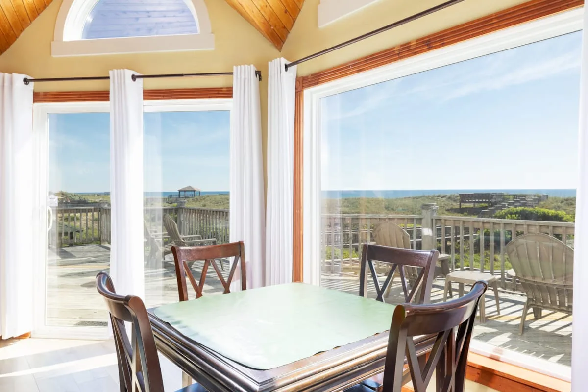 A dining table and chairs sit beside large windows overlooking a deck and the water.