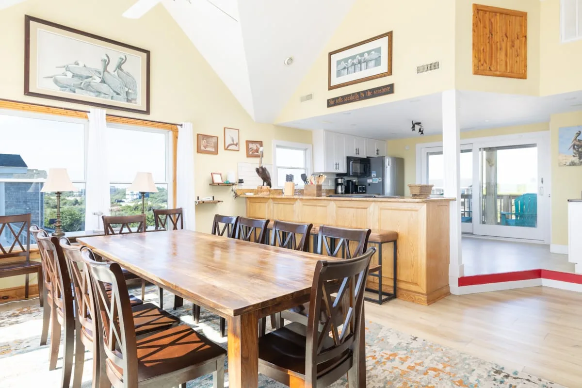 A bright dining area with a long wooden table beside an open kitchen.