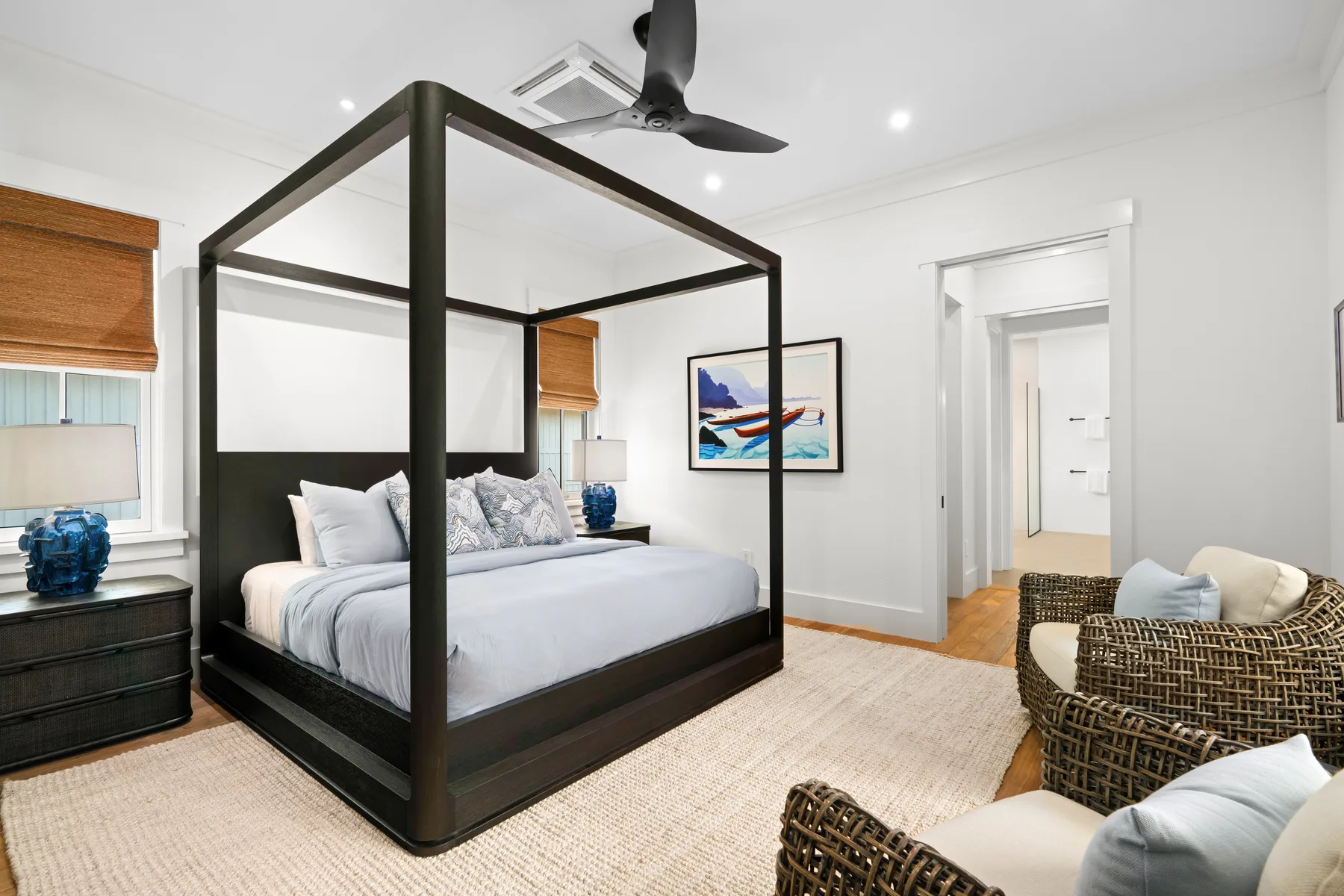 A bright bedroom with a dark four-poster bed and a woven seating area.