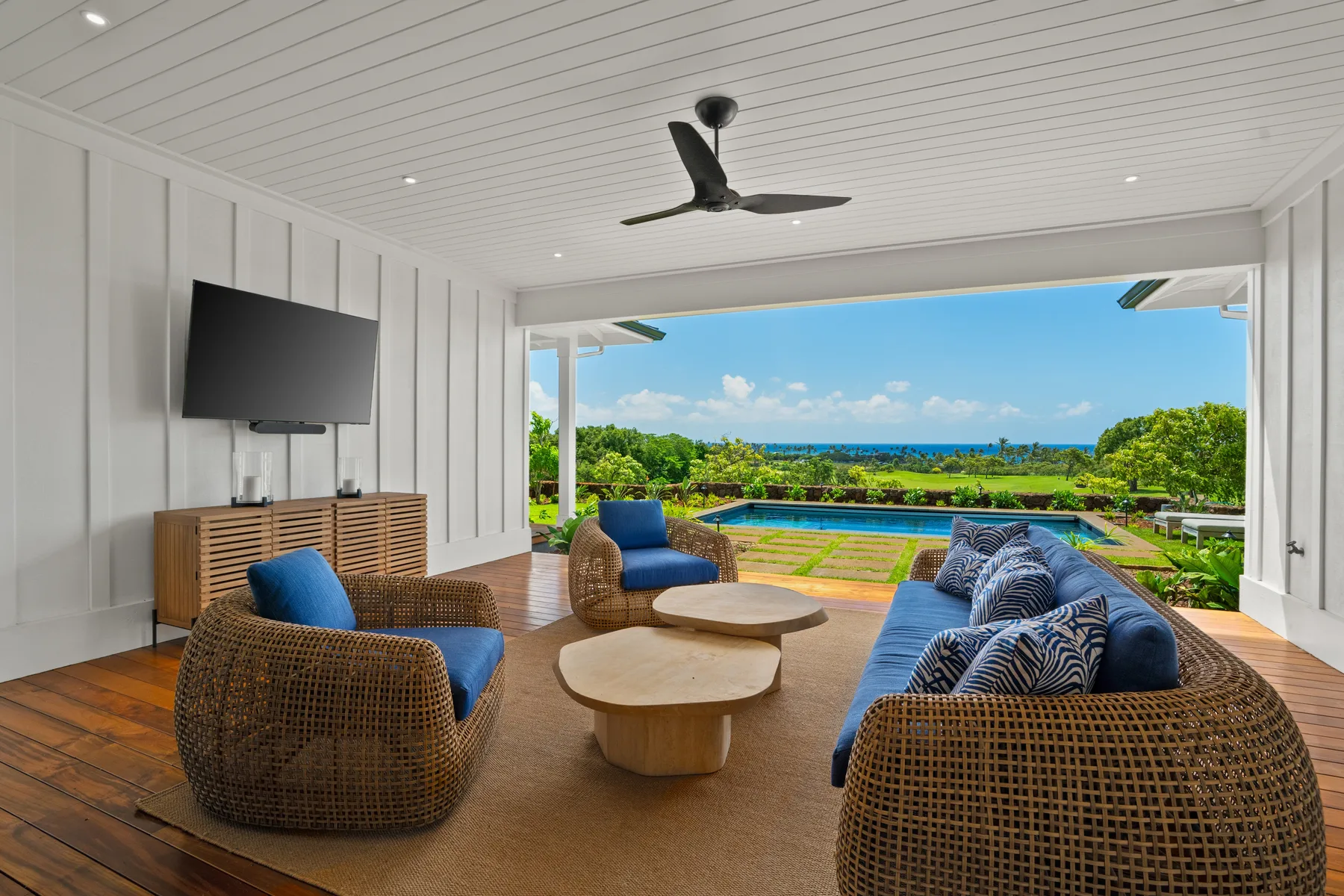 A covered outdoor lounge with wicker seating overlooks a swimming pool and green landscape.