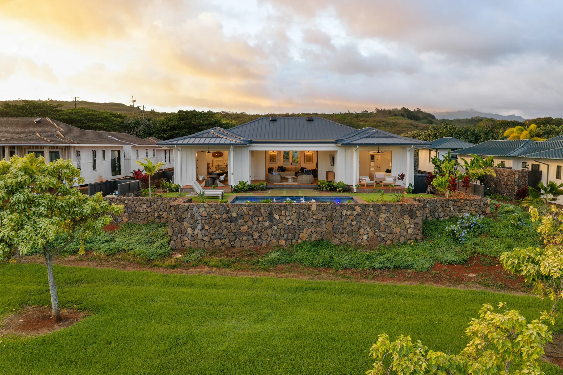 A single-story house with a dark roof sits behind a stone retaining wall and lawn.