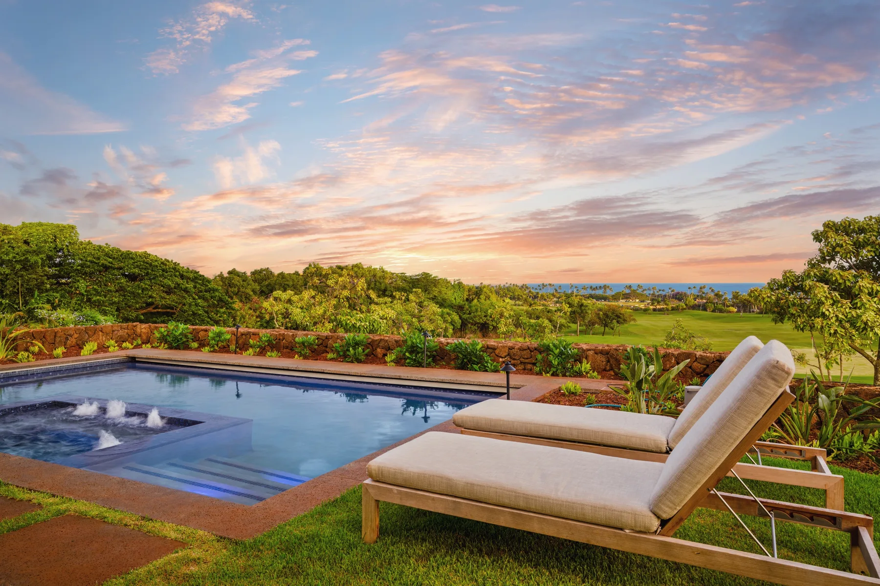 An outdoor pool beside lounge seating overlooks green landscaping at sunset.