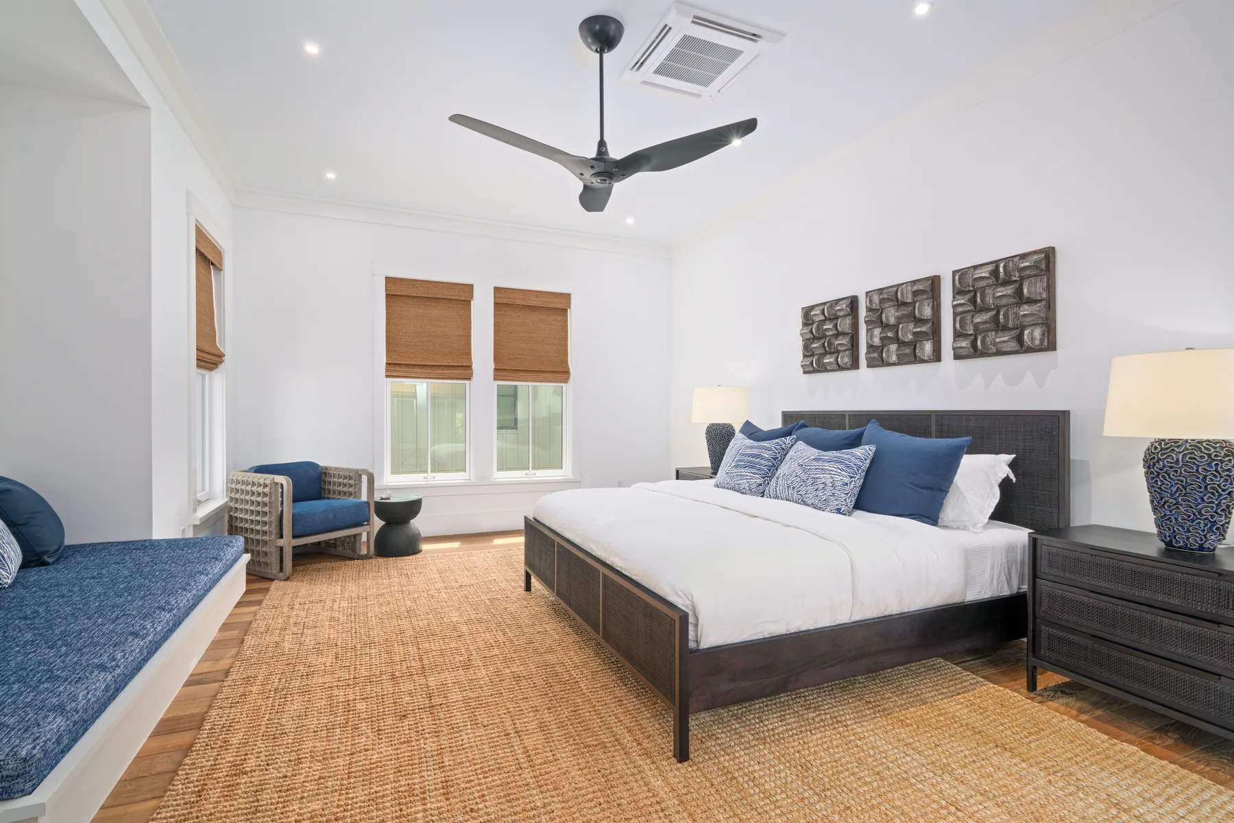 A bright bedroom with a large bed, woven rug, and seating by the windows.