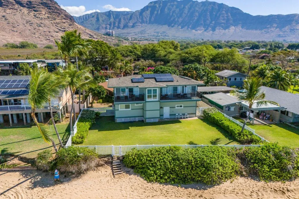 Una casa residencial con un amplio césped, palmeras y montañas al fondo.