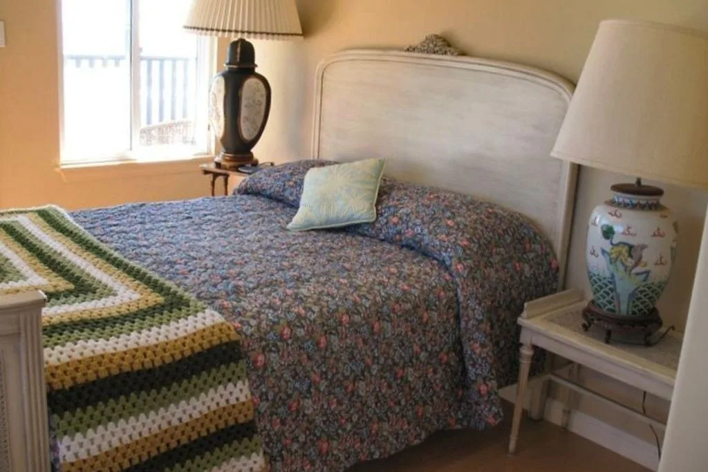 Un dormitorio con una colcha estampada, una ventana y una mesita de noche.