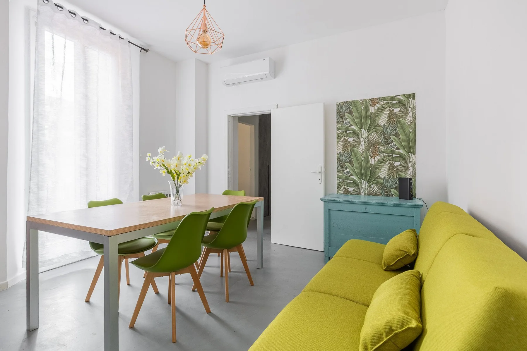 Una luminosa zona de comedor y estar con mesa, asientos verdes y un sofá amarillo.