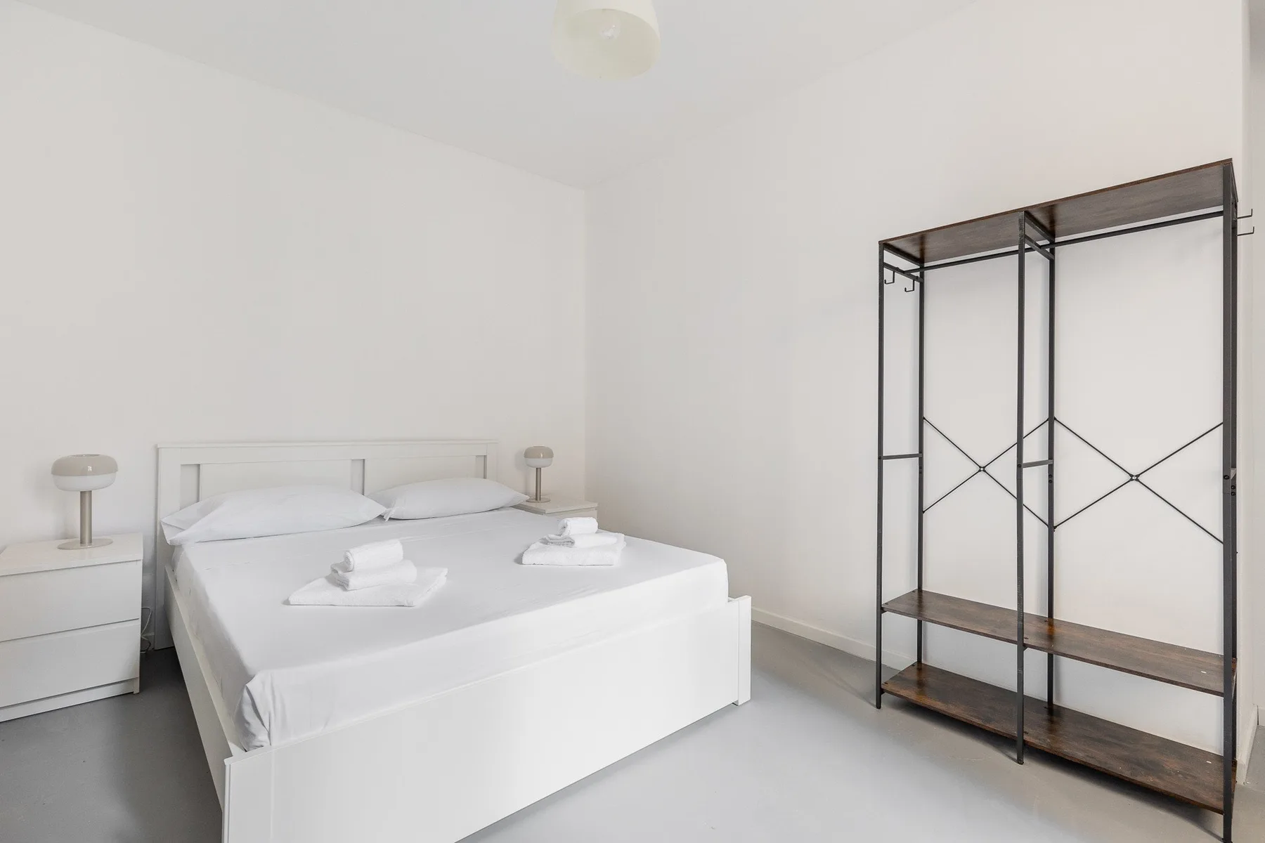 Un dormitorio luminoso con una cama blanca y un perchero metálico abierto.