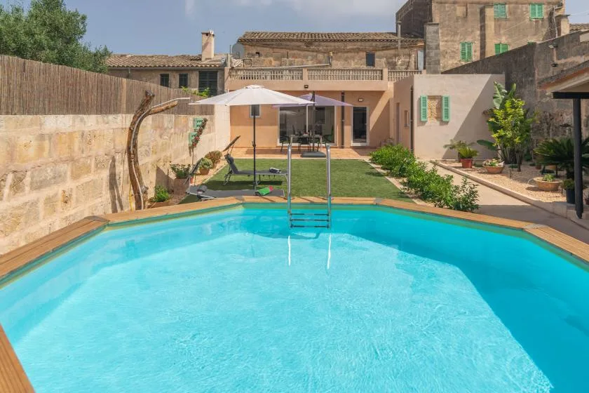 A swimming pool fills the foreground of a sunlit courtyard with a lawn and patio umbrella.
