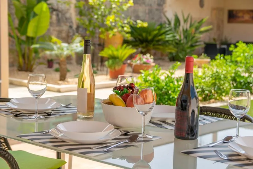 A garden dining table is set with plates, glasses, and a bowl of fruit.