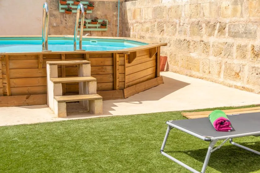 An outdoor pool with wooden sides and steps sits beside a paved patio and lawn.