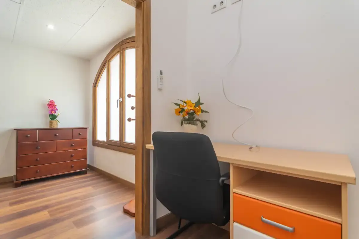 A bright desk and chair sit beside a wooden dresser and arched window.