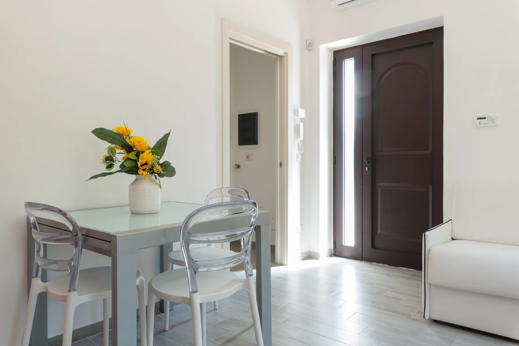 Un comedor luminoso con una mesa de vidrio y una puerta de entrada oscura.