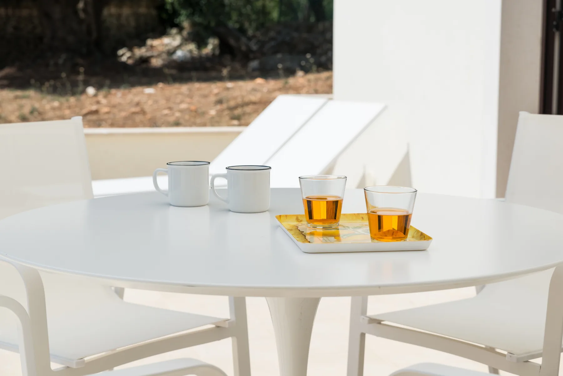 Una mesa blanca de patio sostiene tazas y vasos de té ámbar junto a asientos al aire libre.