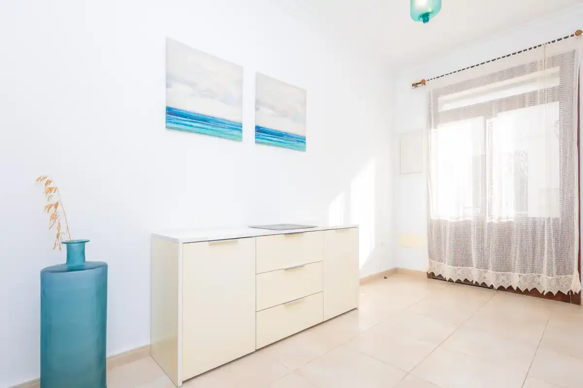 Un dormitorio luminoso con un mueble bajo, dos cuadros de playa y una ventana con cortinas.