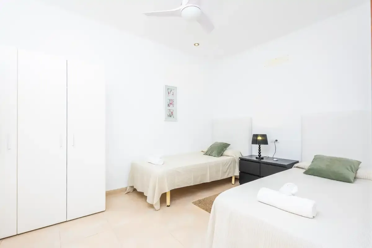 Un dormitorio luminoso con dos camas, una mesita de noche y un ventilador de techo.