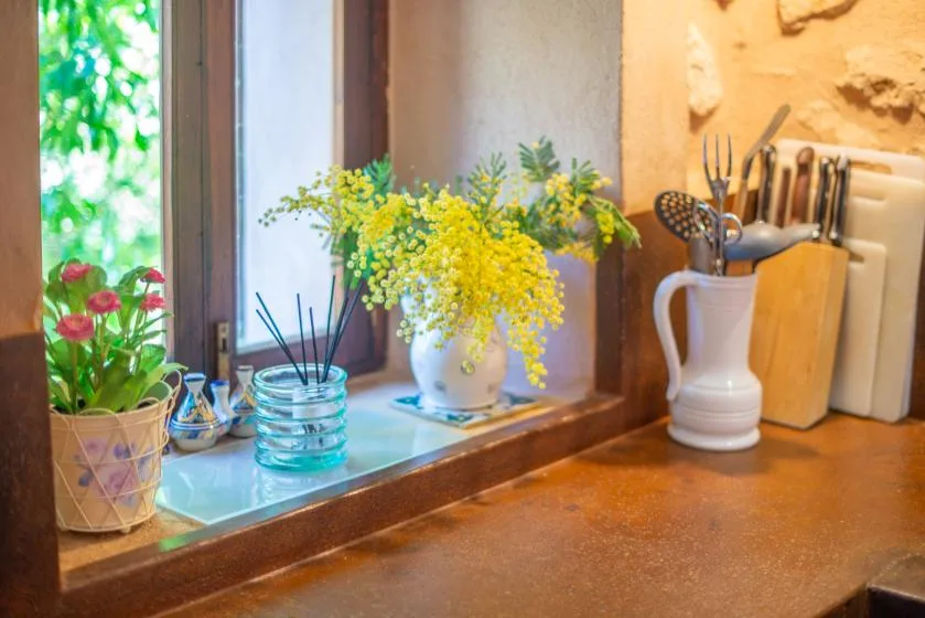 A kitchen counter holds flowers, a reed diffuser, and cooking utensils beside a window.