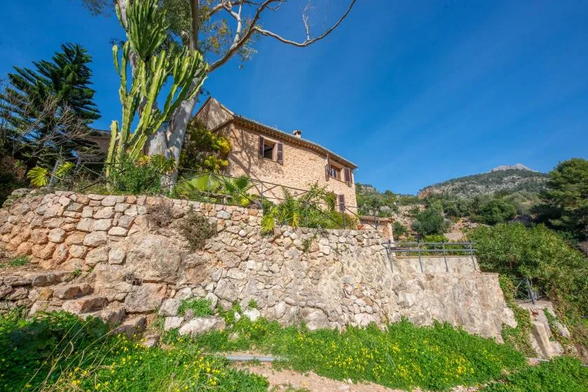 A stone house stands behind a terraced stone retaining wall amid greenery.