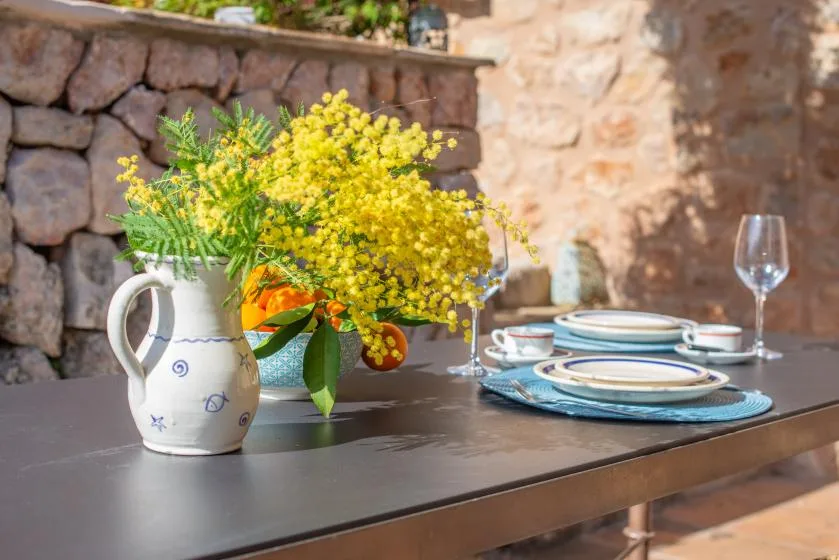 An outdoor dining table is set with plates beside a vase of yellow flowers.