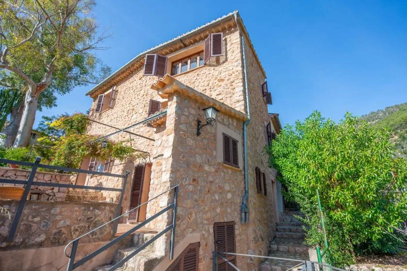 A stone house stands beside a leafy garden and outdoor steps.