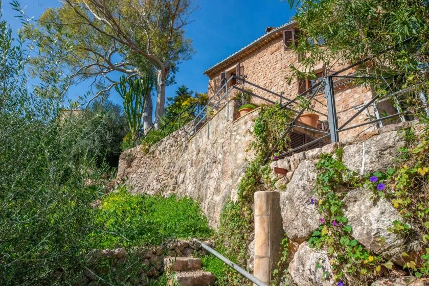 A stone house stands above a terraced garden with steps and greenery.