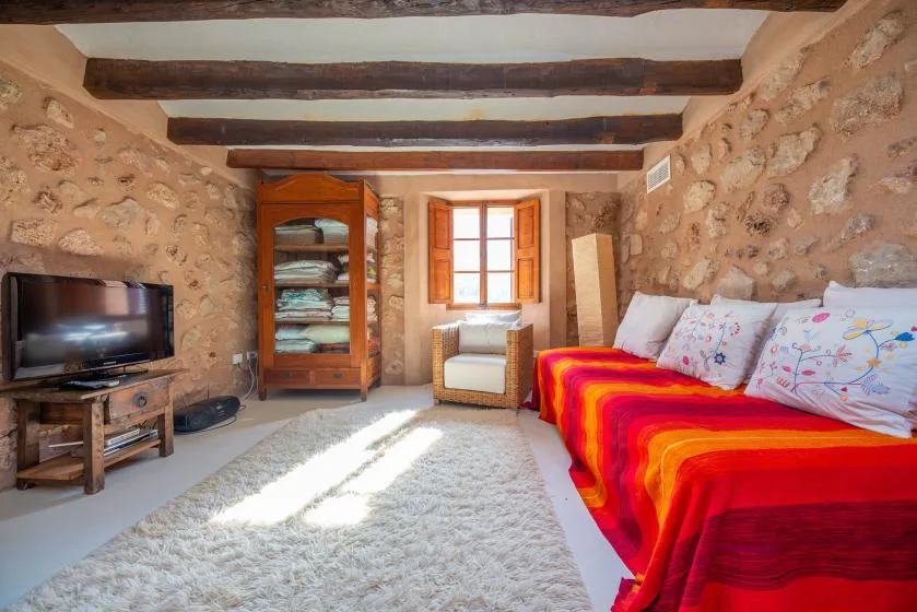 A rustic bedroom with a colorful bed, wooden cabinet, and sunlight through the window.
