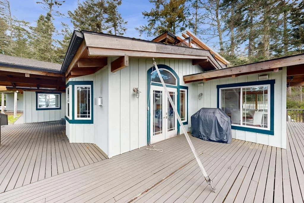 A white house opens onto a broad wooden deck, with trees visible behind it.