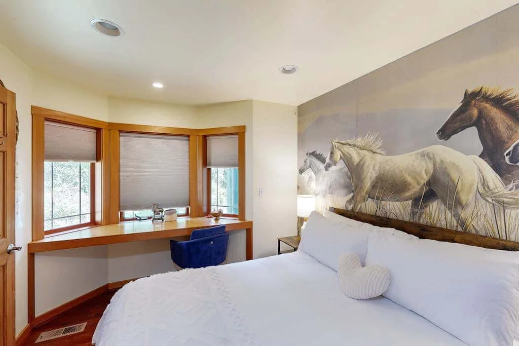 A bedroom with a white bed, a desk by the windows, and a horse mural.