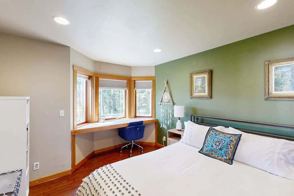 A bedroom with a bed, colorful accent pillow, and a desk beneath the windows.