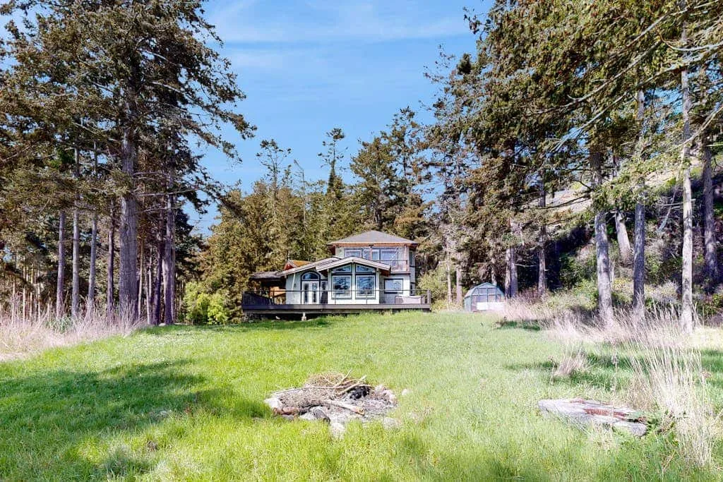 A house stands beyond a grassy clearing, surrounded by tall trees.