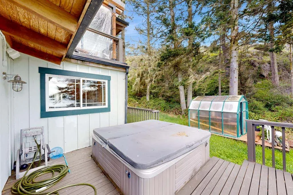 A hot tub sits on a wooden deck beside a small greenhouse and trees.